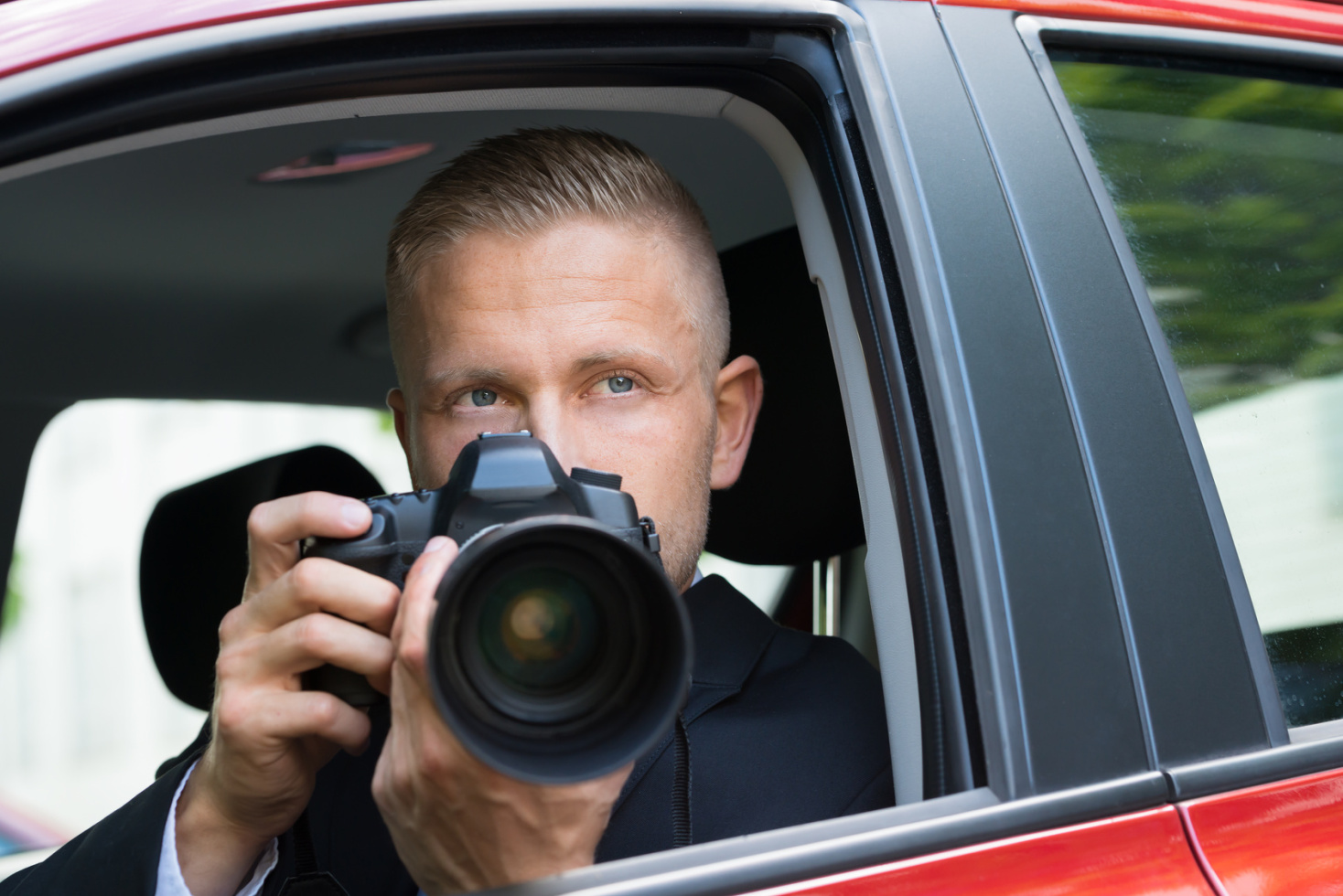 Ein Mann im Anzug sitzt im Auto, blickt durch das offene Fenster nach draußen und hält eine Spiegelreflexkamera in beiden Händen: Wirtschaftsdetekive der Kurtz Detektei Hamburg.