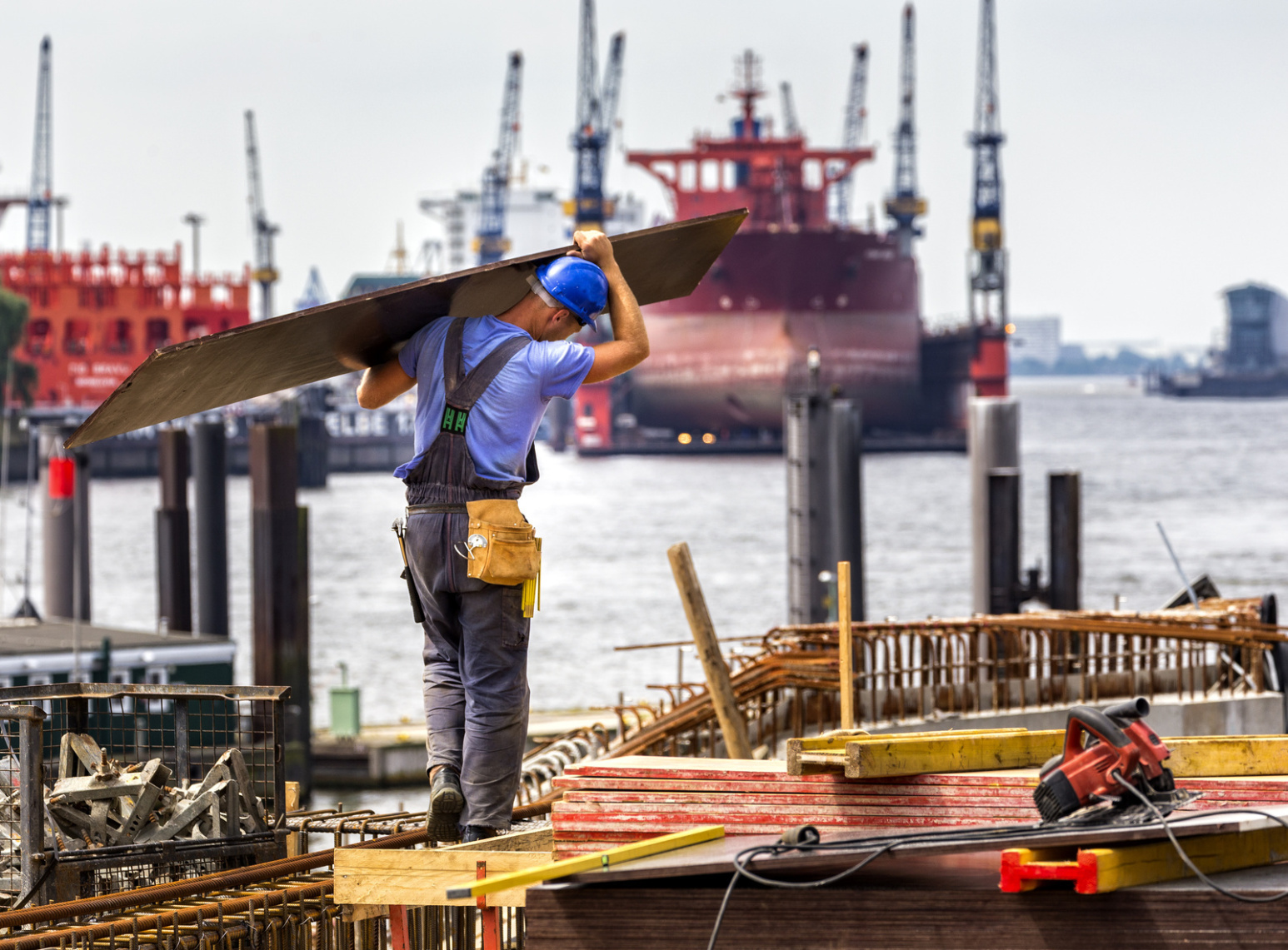 Bauarbeiter schleppt eine Platte im Hamburger Hafen zu einer Baustelle, im Hintergrund Schiffe und Kräne; Kurtz Detektei Hamburg.